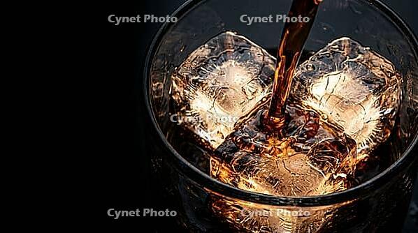 Extreme macro inside short thick-walled rocks glass showing dark rum stream falling from above threading between three large clear craft ice cubes beginning to show hairline fractures from rum contact, single Dedolight spot transmitting through ice turnin [IBR125000326]