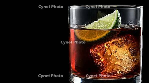 Macro close-up through clear glass wall of short rocks glass containing dark rum with lime wedge resting on large craft ice cube pressing slightly into its surface and dark rum filling to just above lime, Dedolight spot transmitting through glass turning  [IBR125000327]