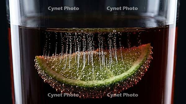 Macro view through clear glass wall of highball glass containing dark rum cola cocktail showing fully submerged lime wedge covered in dense cascading CO2 carbonation bubble trails erupting from every pore as nucleation sites simultaneously, Dedolight back [IBR125000328]
