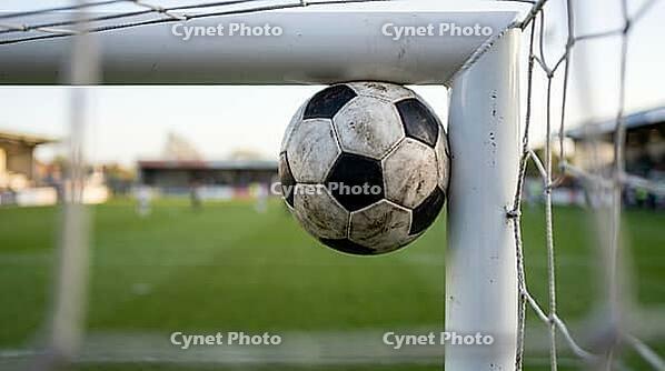 Football wedged in right-angle junction where white goal post meets white crossbar simultaneously touching both metal surfaces, viewed from inside goal looking outward with ball silhouetted against blurred green pitch and pale sky beyond, natural backligh [IBR125000329]