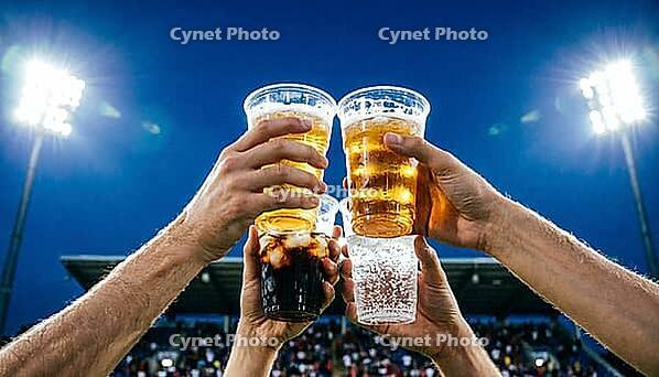 Wide below-angle view looking up at four plastic cups raised simultaneously in diamond formation toast against open night stadium sky, held by four different wrist-level hands with two golden beer one dark cola with ice and one clear sparkling water, stad [IBR125000331]