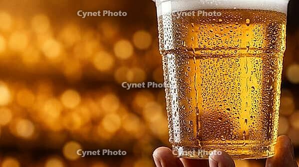 Extreme macro close-up of transparent plastic stadium cup filled with cold golden beer with dense condensation covering exterior surface showing individual droplets and rivulets in sharp detail, single hand visible at base, warm amber stadium bokeh fillin [IBR125000332]