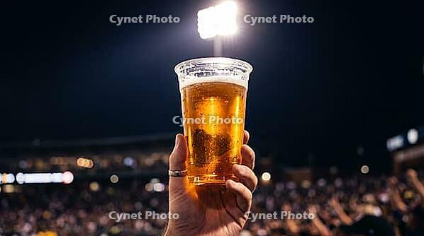 Single transparent plastic stadium cup of golden beer held at arm level against night stadium sky backlit by powerful overhead floodlight creating luminously translucent amber glow through liquid with condensation exterior dots as bright backlit points an [IBR125000333]