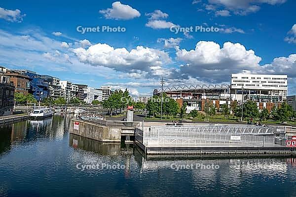 The modern Pole de Commerces et de Loisirs Confluence shopping and leisure center with its curved roof is reflected next to the green promenades of Quai Rambaud in the calm water of the Darse de la Confluence harbour basin under blue summer skies in Lyon, [IBR125002337]