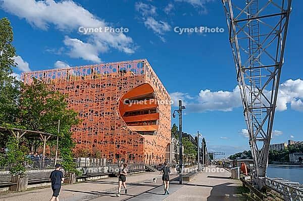 Modern architecture meets industrial history on the banks of the Saone river with the distinctive Le Cube Orange building by architects Jakob and MacFarlane on the Quai Rambaud waterfront with passers-by and a historic crane structure under a blue sky in  [IBR125002338]