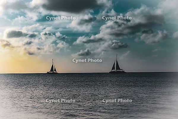 Two sailboats sailing on a tranquil ocean beneath a cloudy sky with a soft sunset glow. Aruba, The Netherlands [IBR125026133]