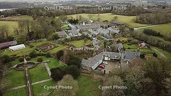 Aerial view of a countryside estate with multiple buildings surrounded by gardens and trees under an overcast sky, Farm in Wales, United Kingdom [IBR125026136]