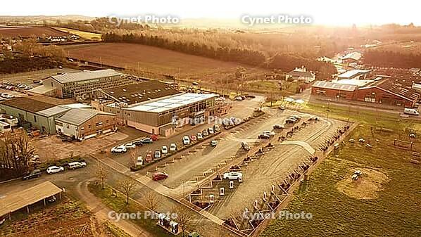 Aerial view of a rural area with buildings, a parking lot, and fields during sunset, Tesla charging station near Exeter, Devon, UK [IBR125026139]
