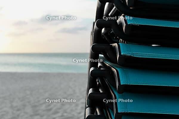 Stacked turquoise beach chairs facing a tranquil ocean and sandy beach during sunset [IBR125026143]