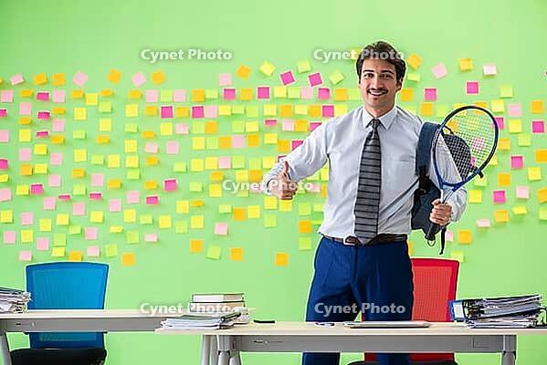 Man with tennis racket in the office with many conflicting priorities [IBR125062163]