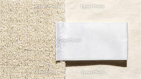 White woven garment care label lying on blended ivory fabric, raking light revealing two distinct thread types in fabric weave below, textile fiber mislabeling and authenticity concept, studio shot, AI generated [IBR125167439]