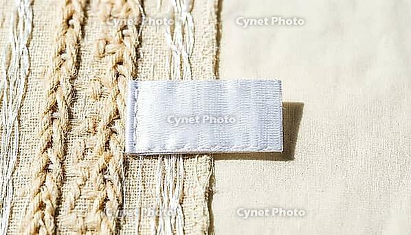 White woven garment care label lying on blended ivory fabric, raking light revealing two distinct thread types in fabric weave below, textile fiber mislabeling and authenticity concept, studio shot, AI generated [IBR125167440]