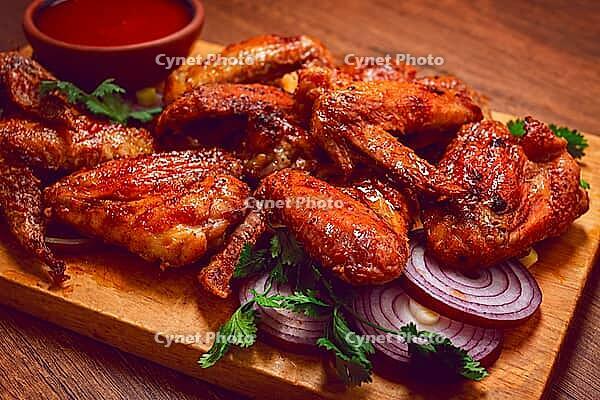 Cooked chicken wings lay on a wooden board. There are slices of onion and cilantro around the wings. A bowl of red sauce is next to the wings for dipping [IBR125177929]