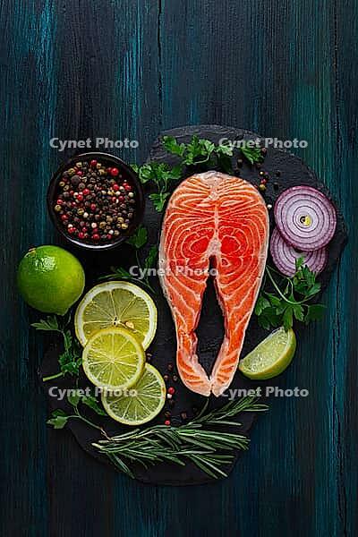 A salmon fillet is placed on a black stone board. Surrounding it are slices of lemon and lime, red onion, herbs, and a bowl of mixed pepper. The setting is on a dark wooden table [IBR125177930]