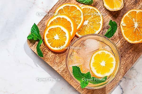 Slices of orange and lemon are arranged on a wooden board next to a glass of drink. Fresh mint leaves add color. The setting looks bright and inviting [IBR125177941]