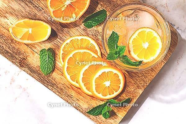 Slices of orange and lemon are arranged on a wooden board next to a glass of drink. Fresh mint leaves add color. The setting looks bright and inviting [IBR125177942]