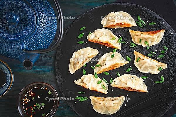 Gyoza, on a black stone platter, with a tetsubin teapot, a bowl with soy sauce, on a wooden table, dark style, no people [IBR125178016]