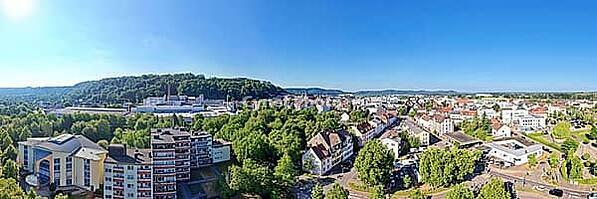A wide-angle panorama from the air shows the city of Homburg on a sunny day. Residential buildings and trees are in the foreground, while the city center with streets and shops runs through the middle. In the background, wooded hills and the distinctive S [IBR125180382]