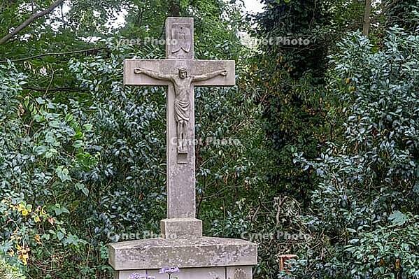 A tall stone cross is placed in a wooded area. Green trees and bushes surround the cross. Small flowers grow at its base. The scene shows nature around the structure during the day, Borken, NRW, Germany [IBR125185853]