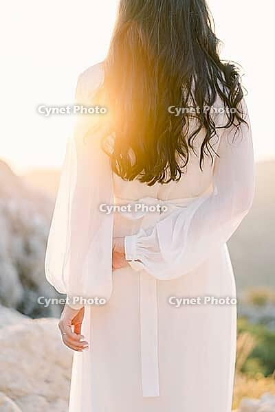 Bride is standing on a mountain touching her other hand with one hand in bright sunlight. Back view. [IBR125188163]