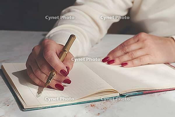 A person writes on an open notebook using a gold pen while sitting at a wooden table. The scene takes place in daylight. The focus is on the writing activity [IBR125204123]