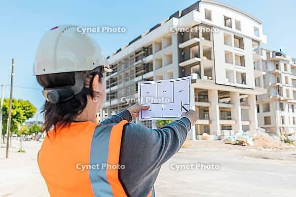 A young Hispanic woman in a hard hat and safety vest examines blueprints at a construction site with a partially built apartment building in the background [IBR125237468]