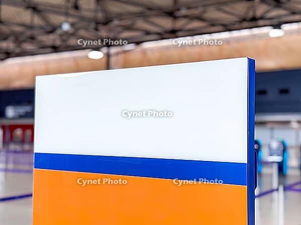 A colorful sign with orange, white, and blue sections stands in an indoor space of airport or terminal. The background features a modern architectural design [IBR125237489]