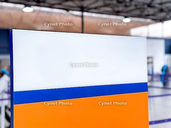 A colorful sign with orange, white, and blue sections stands in an indoor space of airport or terminal. The background features a modern architectural design [IBR125237495]