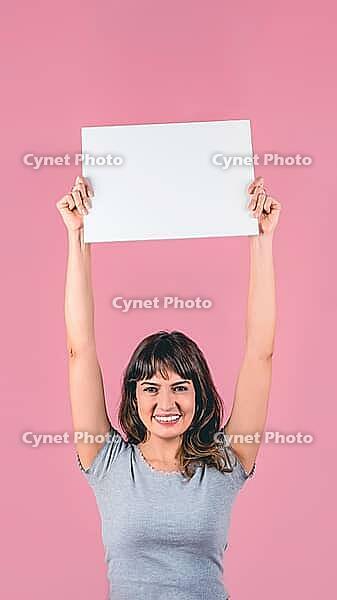 Smiling young woman holding blank sign above head with copy space on pink background [IBR125242401]