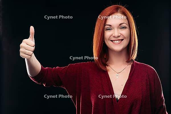 Attractive red haired girl in bordo blouse thinking about something and holding hand with finger up. Young beautiful woman on black background seriously looking at camera. Pretty lady having new idea [IBR125261604]