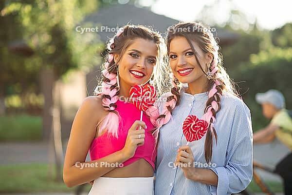 Pretty girlfriends with colorful braids holding candy heart on stick like heart and happy smiling at camera. Gorgeous and stylish women walking at street after shopping and having fun together [IBR125261614]