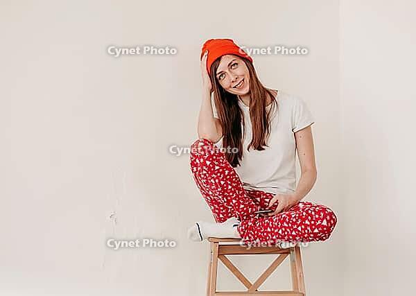Smiling funny girl in a red hat and Christmas pajamas sitting in bed with white sheets. holidays for christmas. Good Christmas mood girl. Girl in a white T-shirt and pajamas, against the background of a white wall, copy space [IBR125261618]