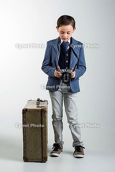 8 year old boy dressed up in suit, standing next to vintage suitcase, looking at photo camera taking up a vintage suitcase. travel concept [IBR125261637]