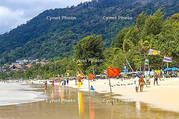 Kathu Phuket Thailand 08. October 2025 Patong Beach bay panorama view with promenade tourists people blue water waves sun loungers parasols lifeguard watch tower mountains and palm trees in Thailand [IBR125280359]