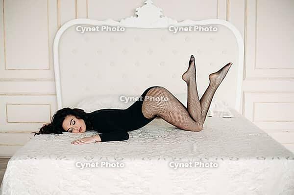 Front view of beautiful and sexy woman in black lying on big bed, looking at camera, holding legs up. Attractive lady with plups lips and posing, looking at camera. Passion and beauty [IBR125280383]