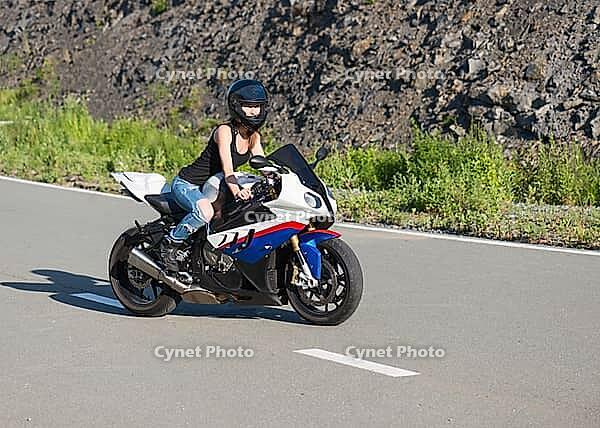 Young beautiful blonde girl in trendy blue jeans and a black T-shirt on the nature behind the wheel of modern motorcycle. Portrait in soft sunny colors [IBR125324100]