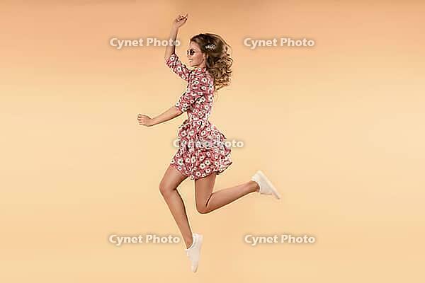Side view of crazy girl wearing pink floral dress and white sneakers jumping and laughing in studio. Attractive young female in glasses enjoying warm weather and stylish outfit. Concept of happiness [IBR125324104]