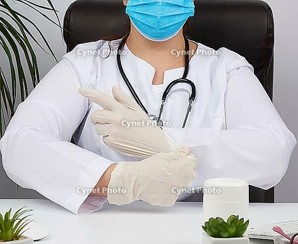 Woman doctor in a white uniform, disposable medical mask is sitting in a chair at the table and puts on his hands white sterile latex gloves, doctor?s office [IBR125331158]