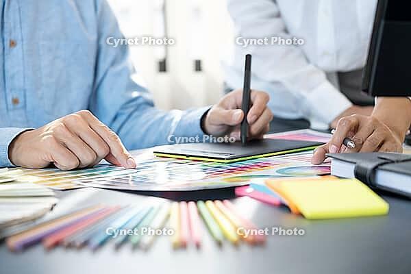Graphic designer or creative working together coloring using graphics tablet and a stylus at desk with colleague [IBR125331173]
