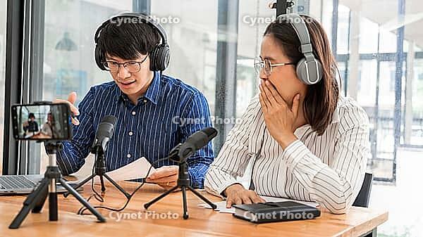 Asian Man and woman podcasters in headphones recording content with colleague talking to microphone and camera in broadcast studio together, communication technology and entertainment concept [IBR125331186]