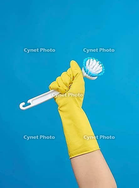 Hand in a yellow rubber glove for cleaning a house holds plastic brush with a handle on a blue background [IBR125344241]