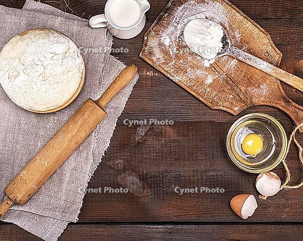 Yeast dough in a wooden bowl on a brown table and ingredients, top view, empty space in the middle [IBR125344263]