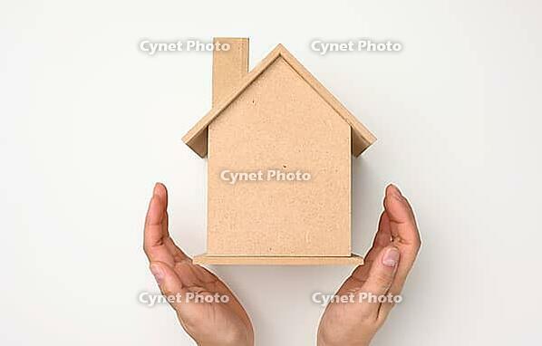 Female hands folded to each other over a wooden miniature model house on a white background. Real estate insurance concept, environmental protection, family happiness [IBR125344299]