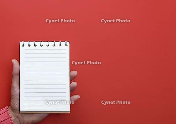 Female hand holding an open notebook with blank white pages on a red background, copy space [IBR125344301]