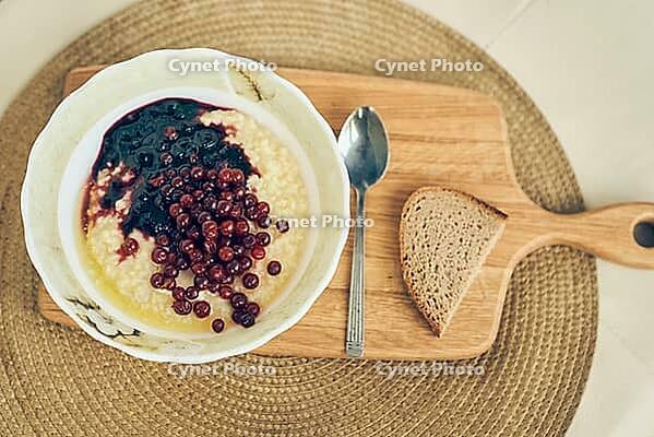 Top view of a plate of wheat porridge with red currants and blueberry jam. A plate of wheat porridge for a healthy gluten-free breakfast, next to a spoon and a piece of bread. [IBR125344317]