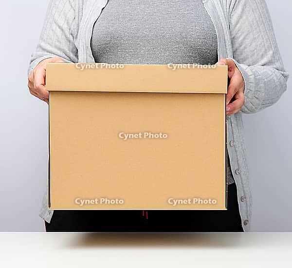 Woman in gray clothes stands and holds a brown box on a white background, moving, sending and delivering goods [IBR125344330]