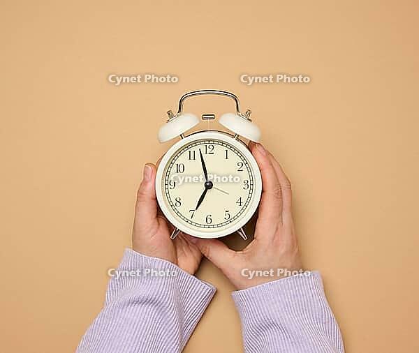 Two female hands hold a round alarm clock, the time is seven in the morning [IBR125363541]