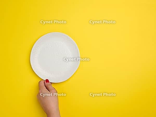 Female hand holding a white empty round plate on a yellow background, top view, copy space [IBR125363576]