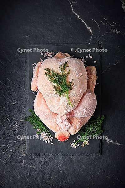 A top view of a whole frozen chicken on a black slate with dill and pink salt, vertical shot. The raw poultry meat, which is covered with frost, stands against a dark stone background [IBR125363613]