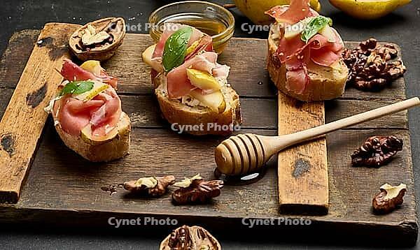 Italian bruschetta with prosciutto, pear slices, cream cheese and fresh basil, garnished with sesame seeds, served on a rustic wooden board [IBR125382560]